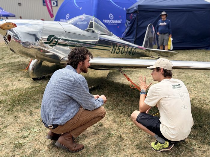 Nate crouched beside a polished vintage aircraft at EAA AirVenture Oshkosh, talking with a fellow engineer wearing a Spirit Engineering Inc. shirt